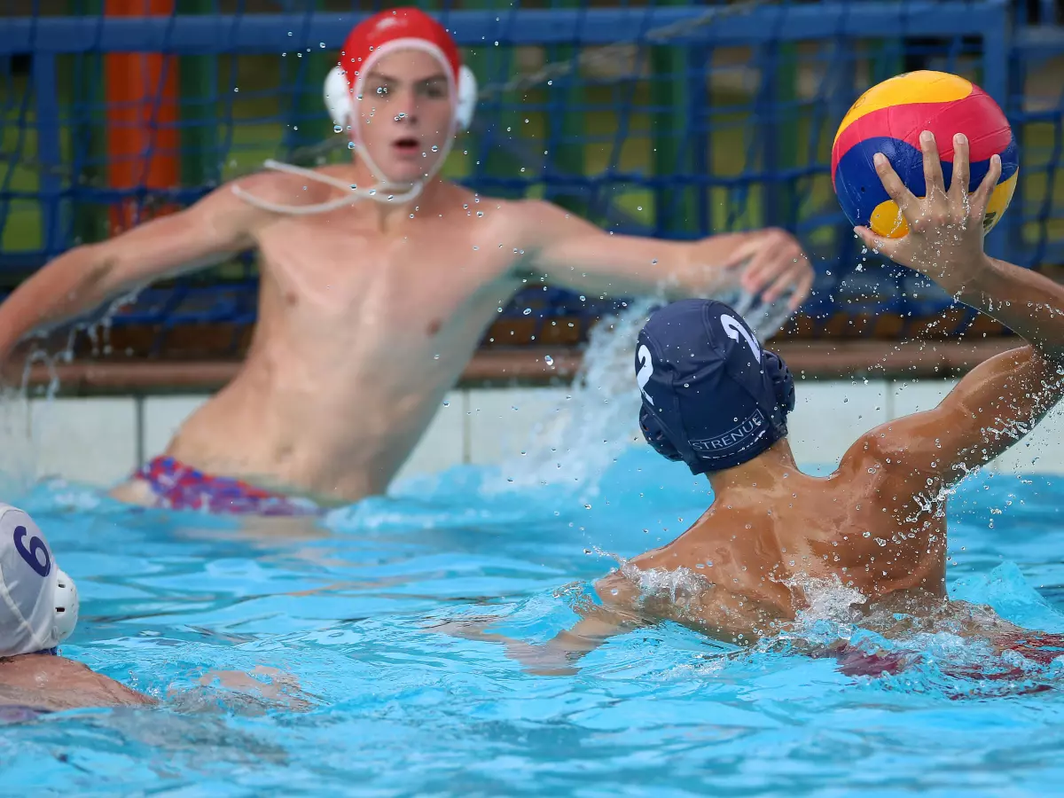 Two water polo players are in the pool with the ball. The player in the foreground is wearing a white swim cap.