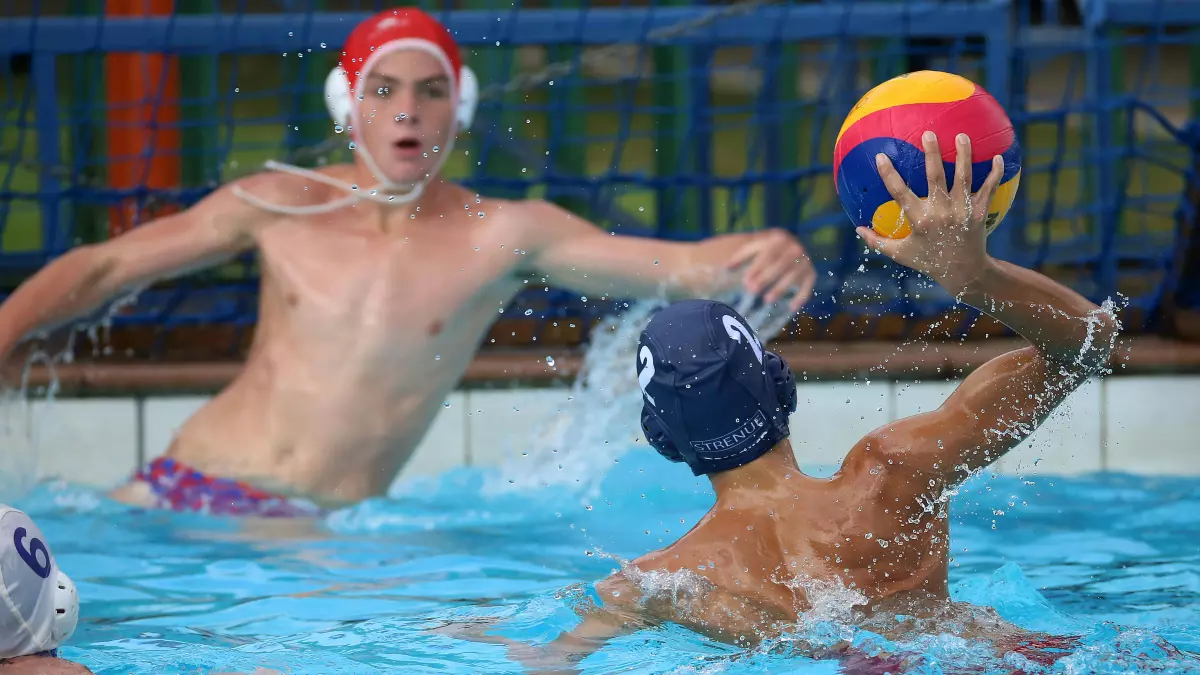 Two water polo players are in the pool with the ball. The player in the foreground is wearing a white swim cap.