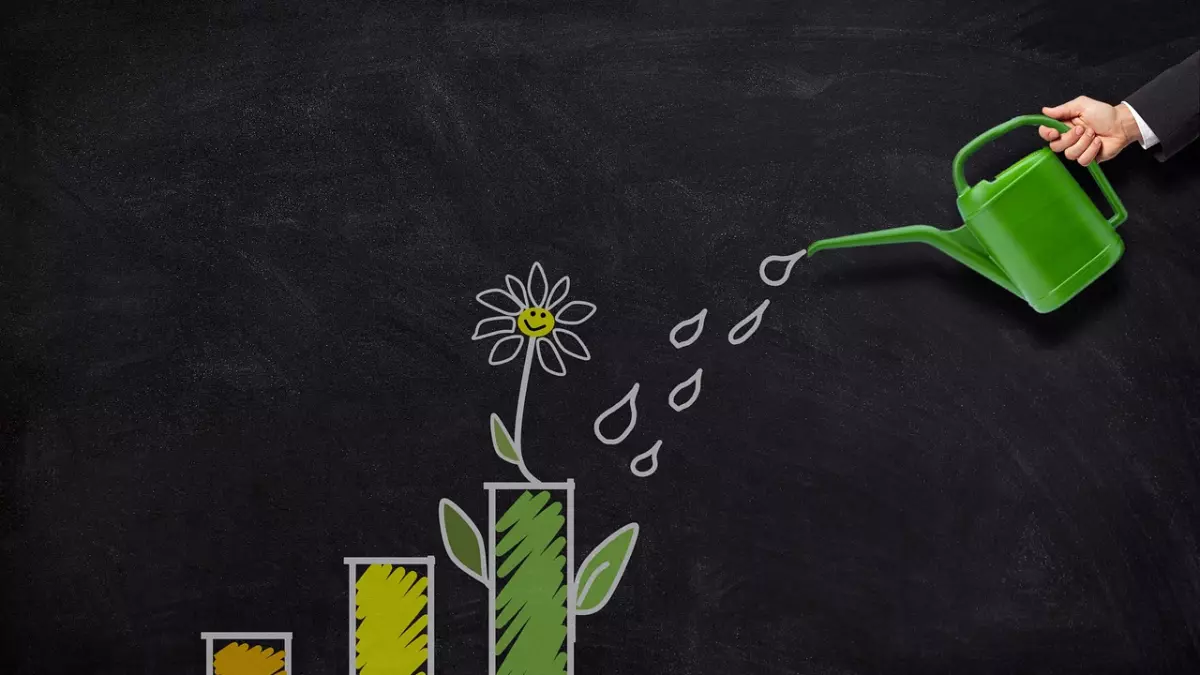 A hand holding a green watering can pouring water onto a flower growing from a bar graph drawn on a blackboard.