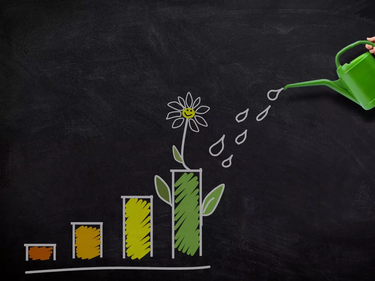 A hand holding a green watering can pouring water onto a flower growing from a bar graph drawn on a blackboard.