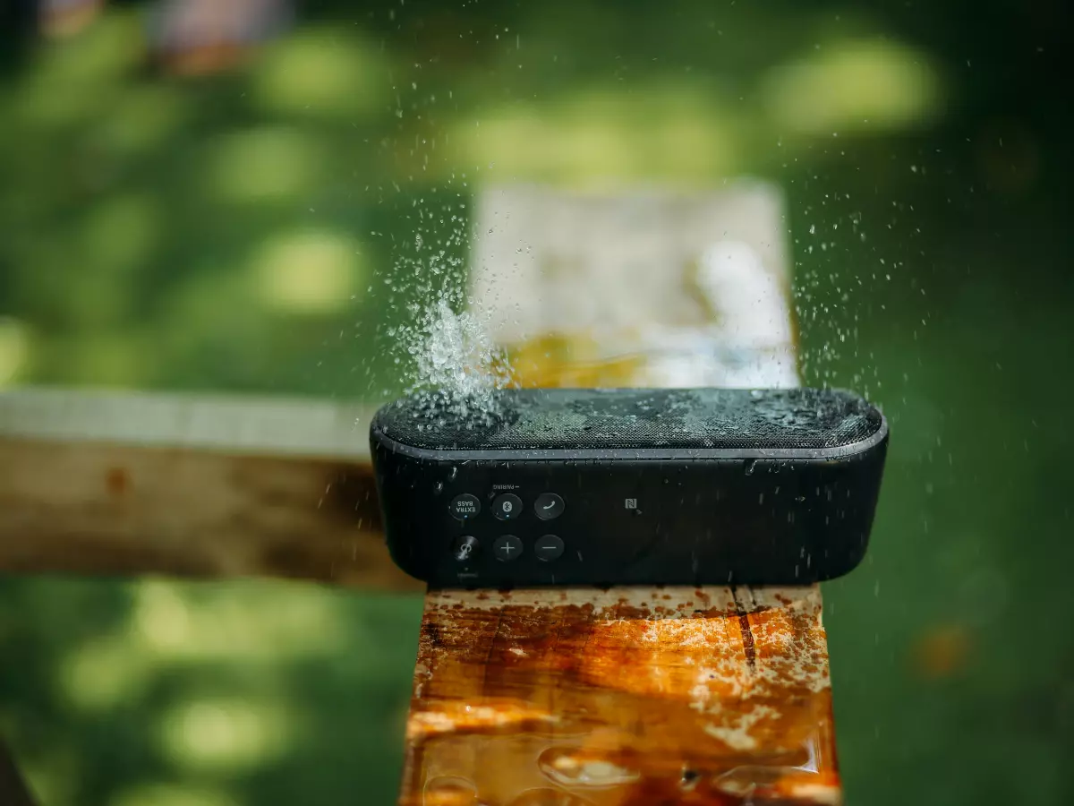 A black Bluetooth speaker is sitting on a wooden surface with water droplets falling on it.  