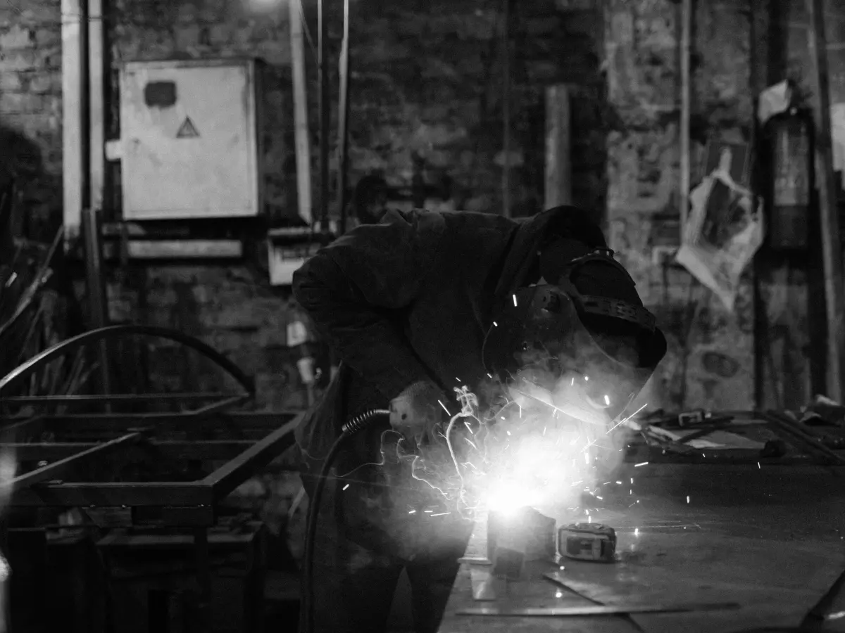 A worker wearing protective gear welds a metal object in a dimly lit industrial setting.  Sparks fly from the weld.