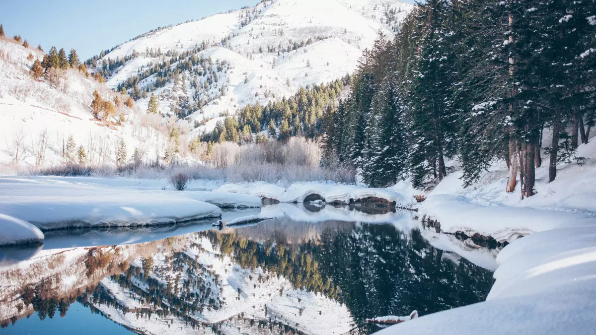 A picturesque winter landscape featuring a snow-covered river reflecting the surrounding snowy mountains and evergreen trees.