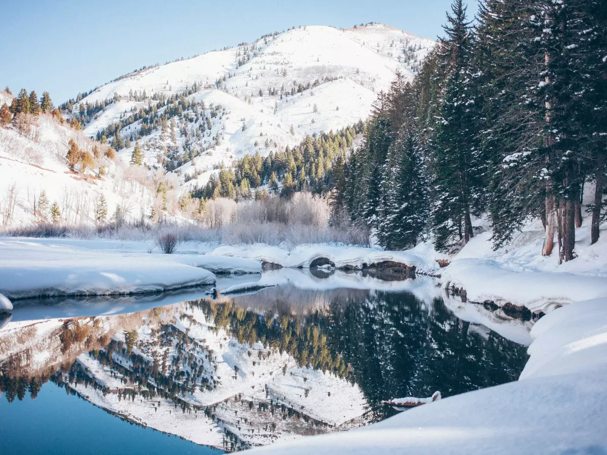 A picturesque winter landscape featuring a snow-covered river reflecting the surrounding snowy mountains and evergreen trees.