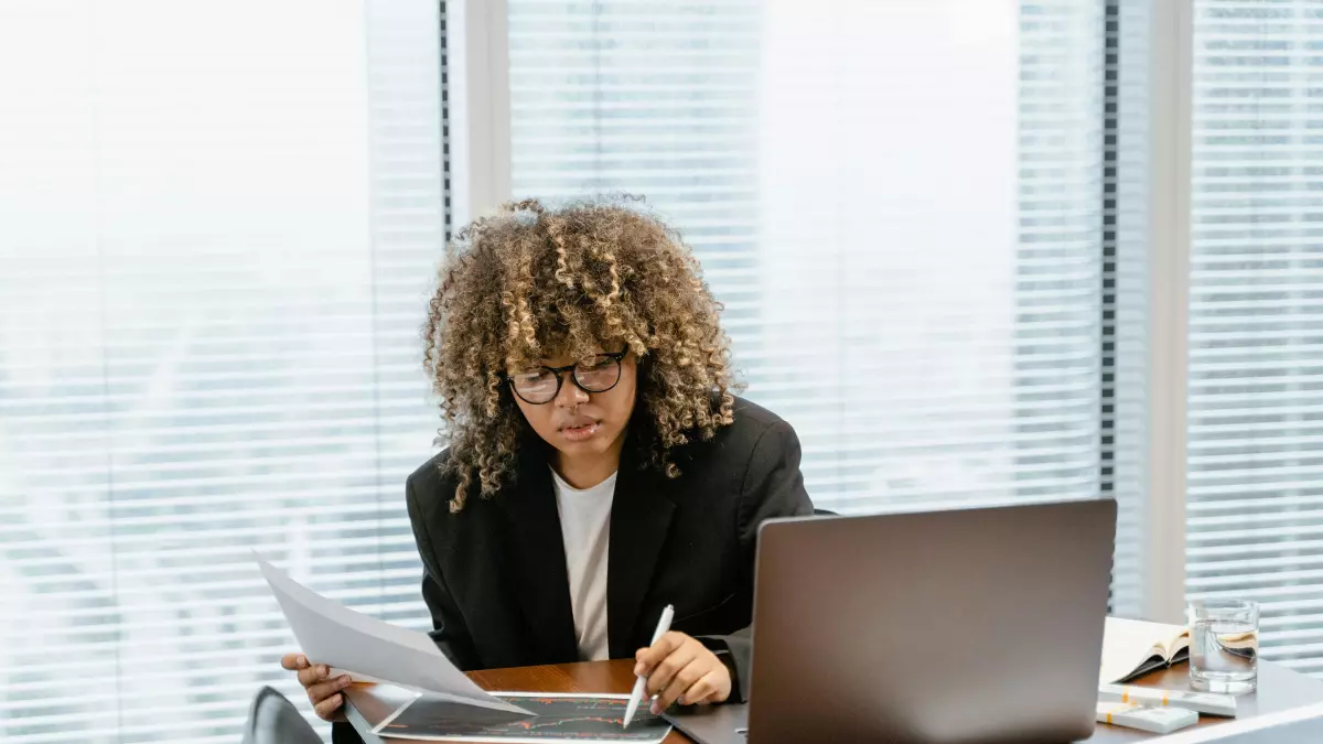 A woman with curly hair is sitting at a desk in front of a window. She is wearing a black blazer and a white shirt. She is looking at a document in her hand, and she is writing on a tablet with a stylus. A laptop is open in front of her on the desk.