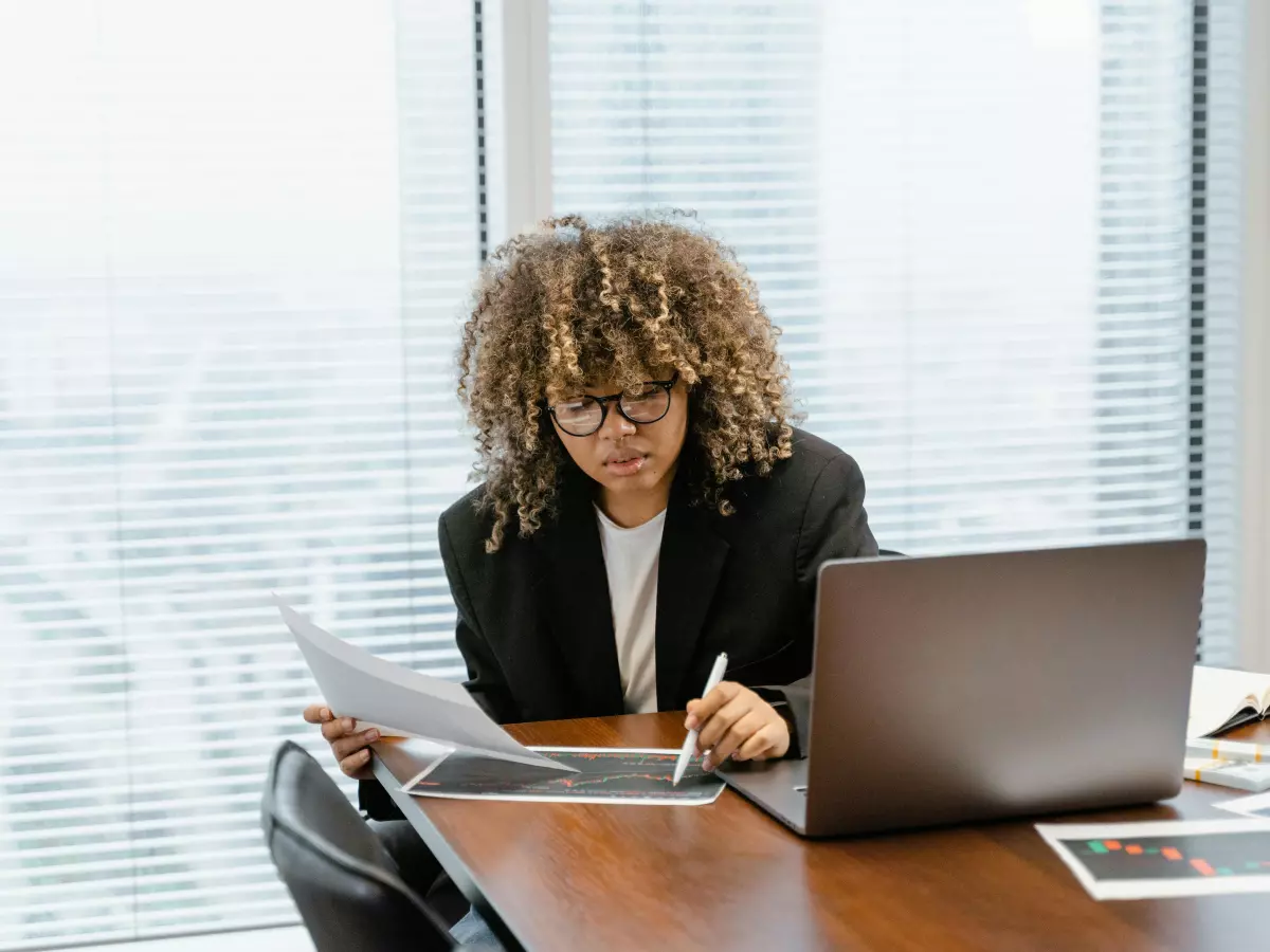 A woman with curly hair is sitting at a desk in front of a window. She is wearing a black blazer and a white shirt. She is looking at a document in her hand, and she is writing on a tablet with a stylus. A laptop is open in front of her on the desk.