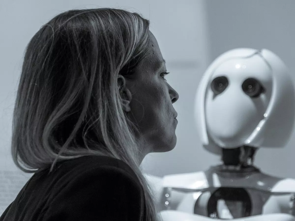 A woman looking intently at a humanoid robot, both in a white room, with the robot's eyes clearly visible.