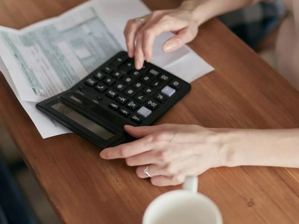 A person's hands using a calculator and a paper on a wooden table, a cup of coffee is in the background.