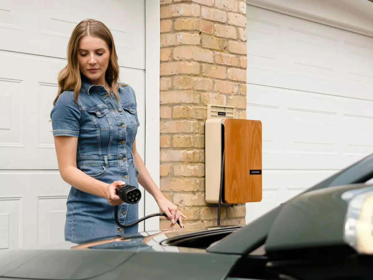 A young woman in a blue jean dress and sneakers is standing in front of a white garage with two double doors, she holds a charging cable that is plugged into an EV car.