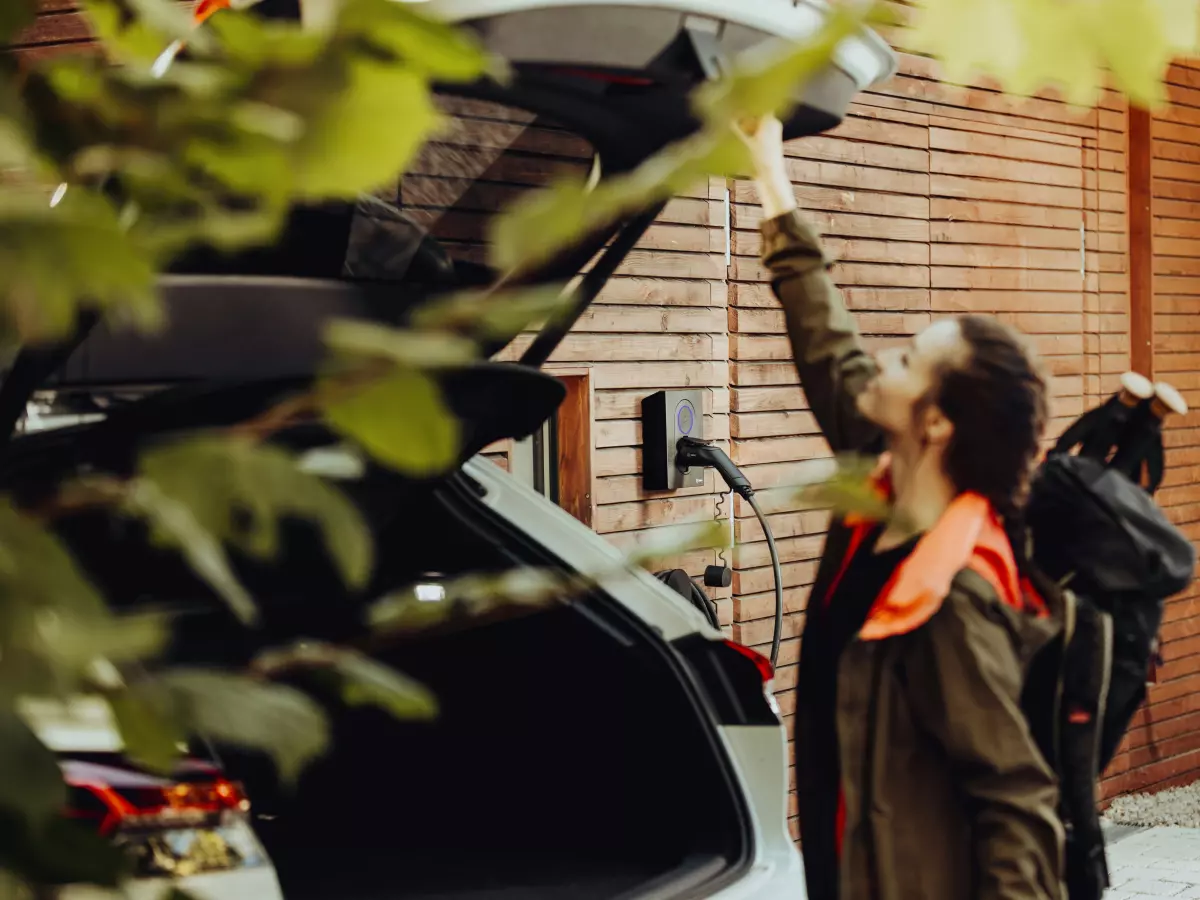 A woman is opening the trunk of an electric car and plugging it into a charging station.