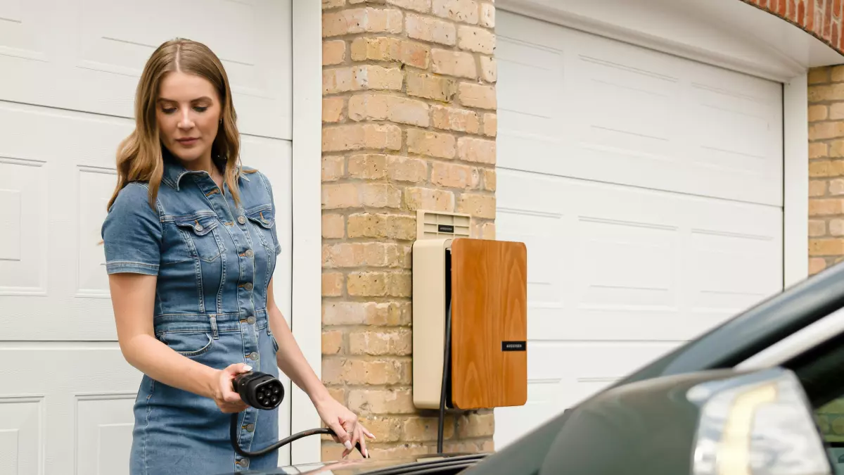 A young woman in a blue jean dress and sneakers is standing in front of a white garage with two double doors, she holds a charging cable that is plugged into an EV car.