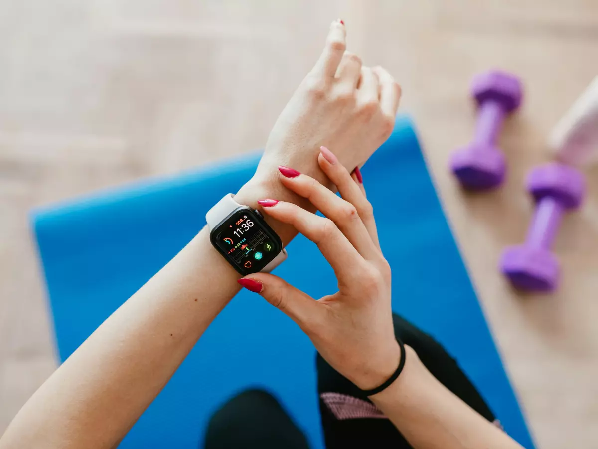 A woman's hand wearing a white smartwatch is on a blue yoga mat. The hand is touching the screen. Two purple dumbbells and a white bottle of water are on the mat.