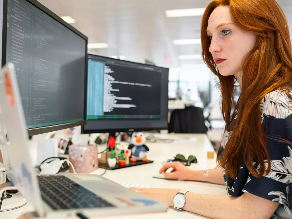 A woman is working on a laptop and looking at the screen with a serious expression. She is sitting at a desk in an office with a computer monitor next to her. She is wearing a black and white patterned shirt and a watch.