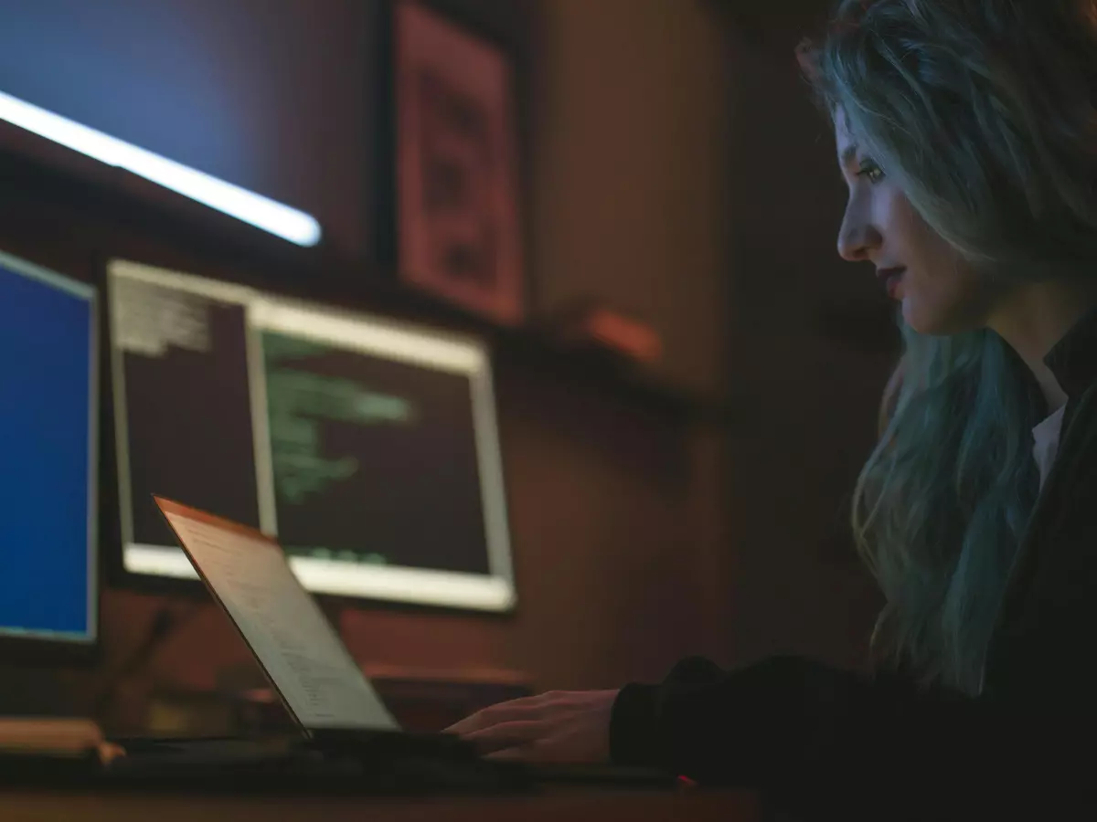 A person in a hoodie sits at a desk in front of a computer. The person is typing on a keyboard, and their face is obscured by the hood of their sweatshirt. The image has a dark and mysterious tone, and the person's focus on the computer suggests they are engaged in a serious task. The image is well-lit, and the background includes a variety of items, including multiple monitors and other computer equipment. The image is reminiscent of a scene from a spy thriller, and it evokes a sense of tension and suspense.