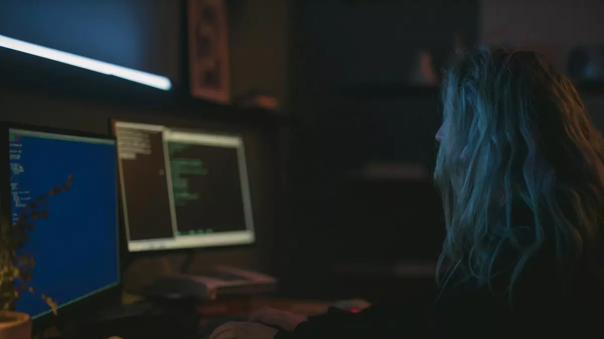 A woman with blue hair is sitting in front of a computer, coding.  The computer screen is displaying a lot of text, likely code, with the background showing another computer screen.