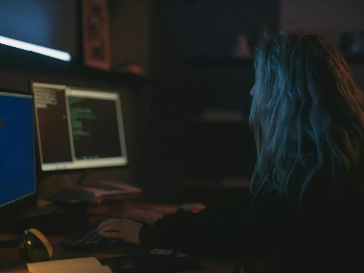 A woman with blue hair is sitting in front of a computer, coding.  The computer screen is displaying a lot of text, likely code, with the background showing another computer screen.