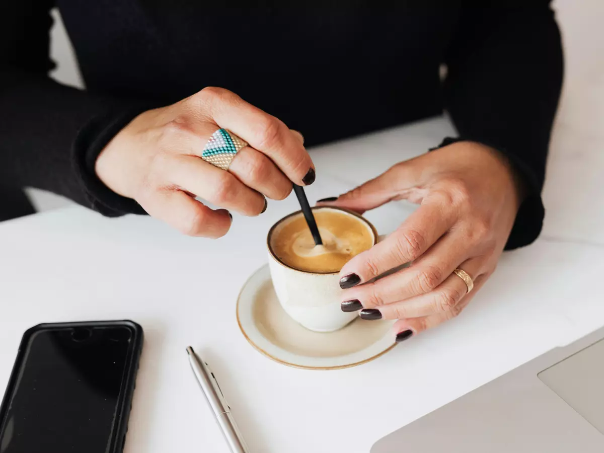 A woman's hand wearing a blue smart ring on her index finger. She is stirring her coffee with a black straw. Her black nail polish is visible. 