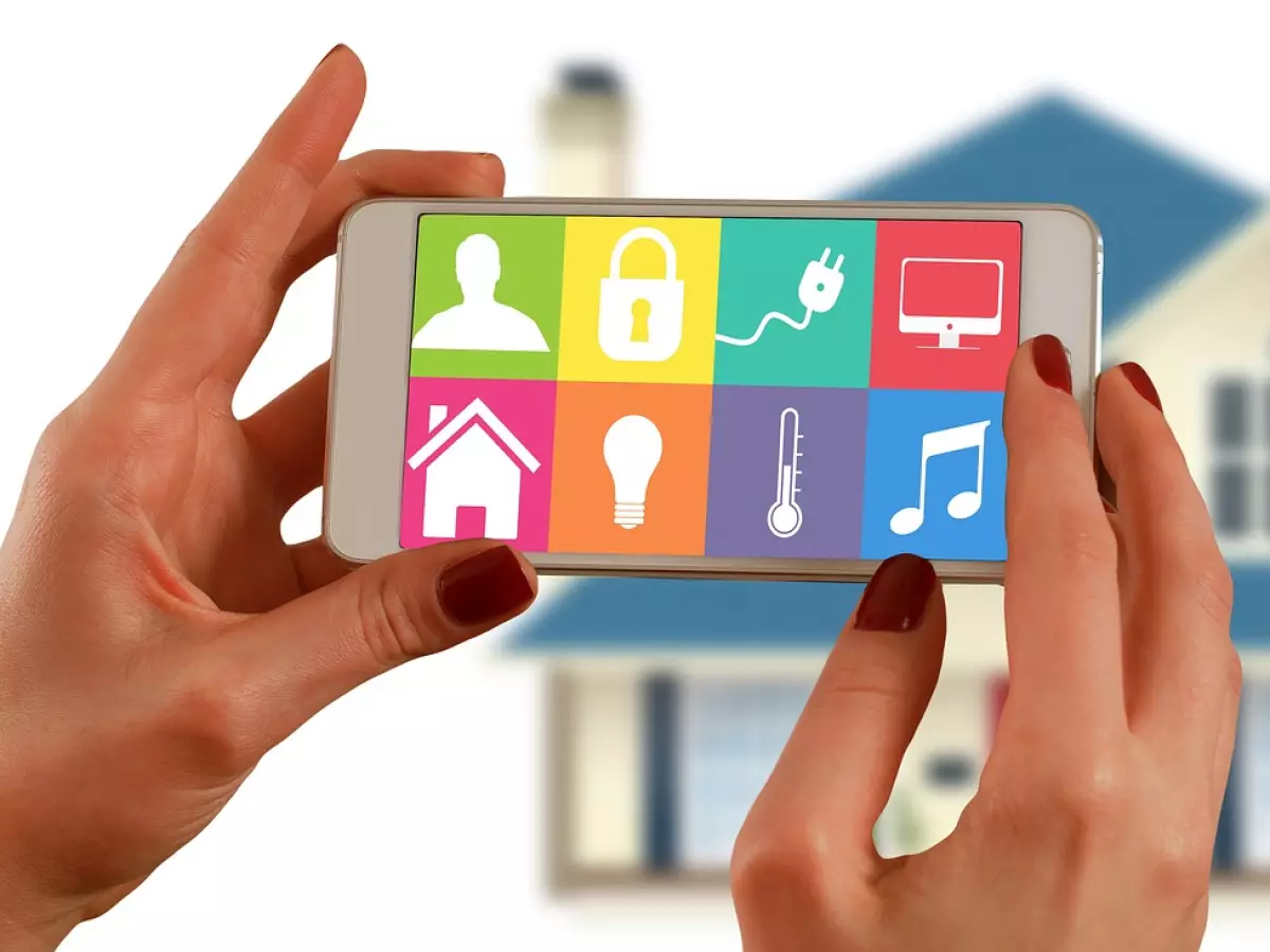 A woman's hands hold a smartphone displaying various app icons related to smart home functions. A blurred image of a house is visible in the background.