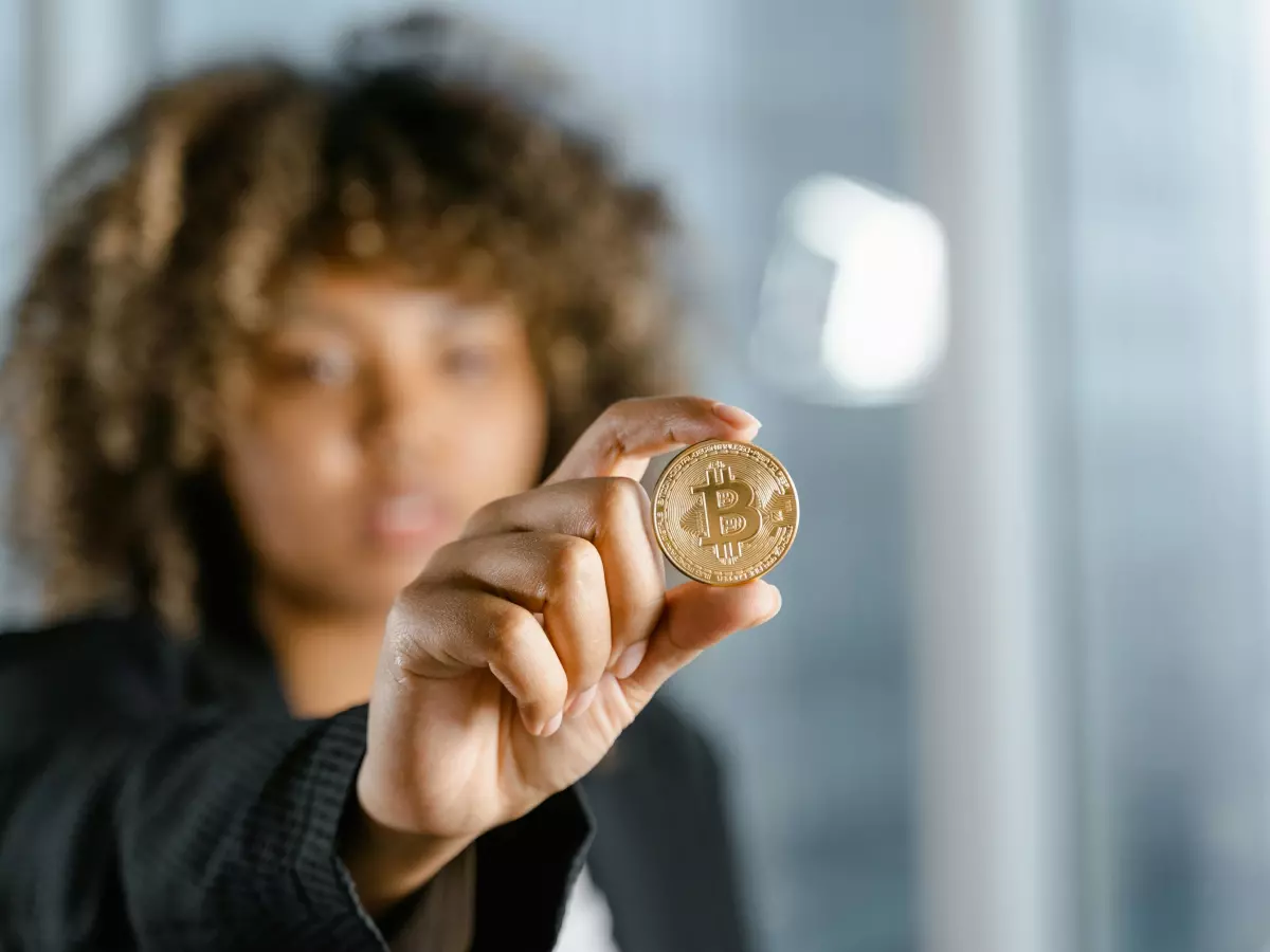 A young woman holding a Bitcoin in her hand, looking at the camera with a serious expression. The background is blurry and out of focus.