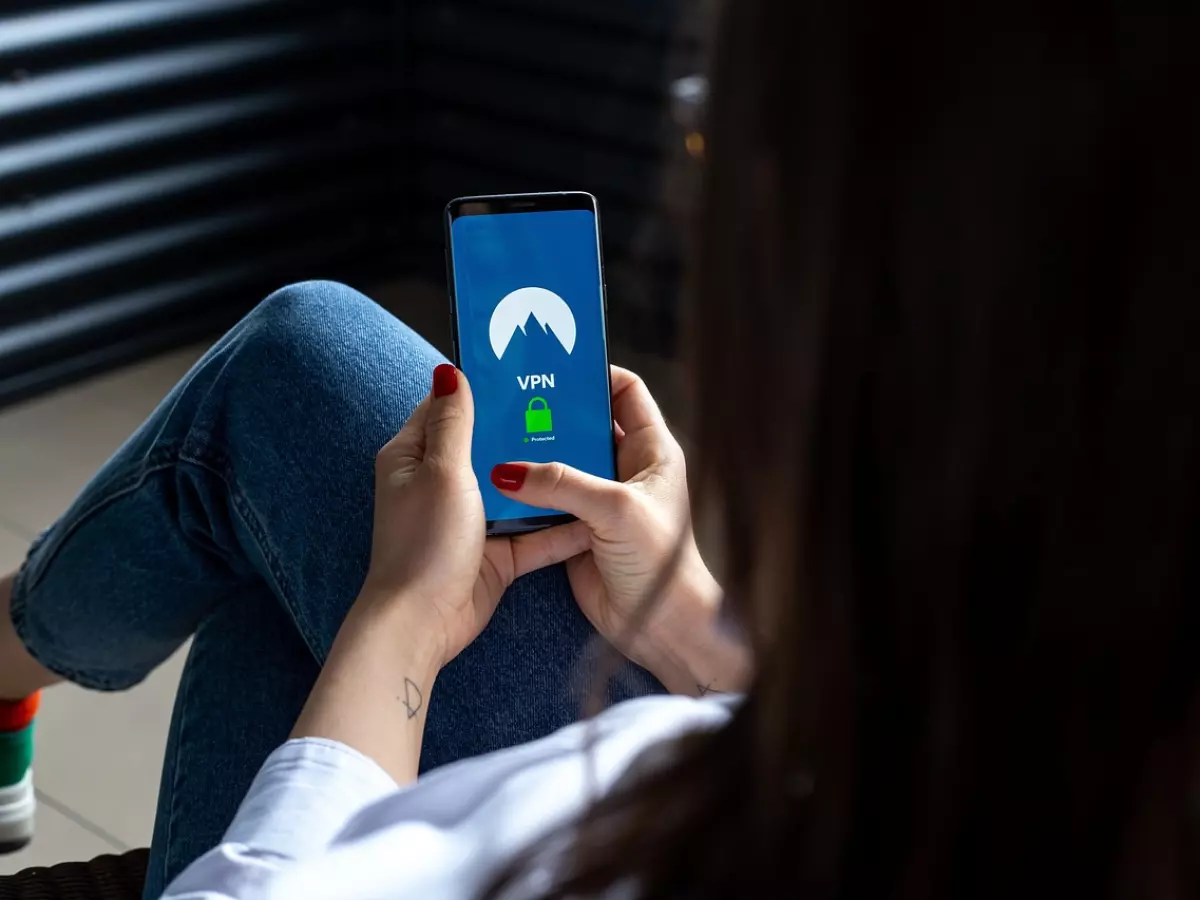 A woman's hands holding a smartphone with a VPN app open on the screen. The image is shot from behind the woman, so her face is not visible. The woman is wearing jeans and a white shirt. The background is blurry and out of focus.