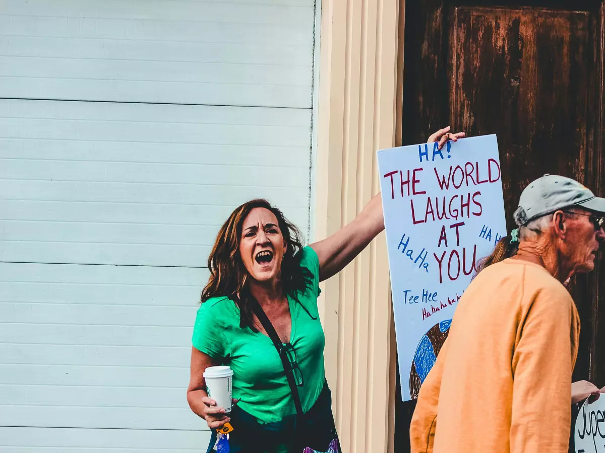 A woman is holding a sign that says "MAY THE WORLD LAUGH AT YOU" and she is yelling, a man is standing next to her, they are both visible, the background is out of focus