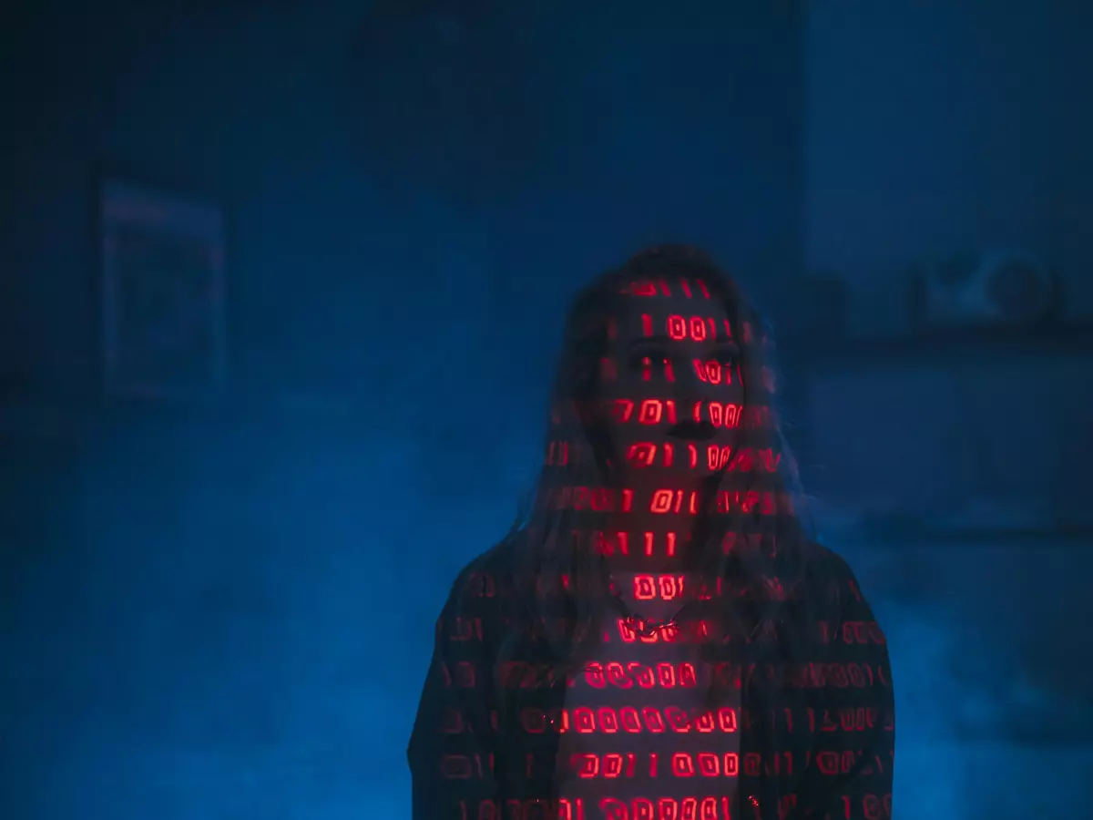 A person in a hooded jacket stands in a dimly lit room, their face obscured by a glowing red code that appears to be forming a digital mask. Blue smoke or mist fills the background, creating an eerie atmosphere. 