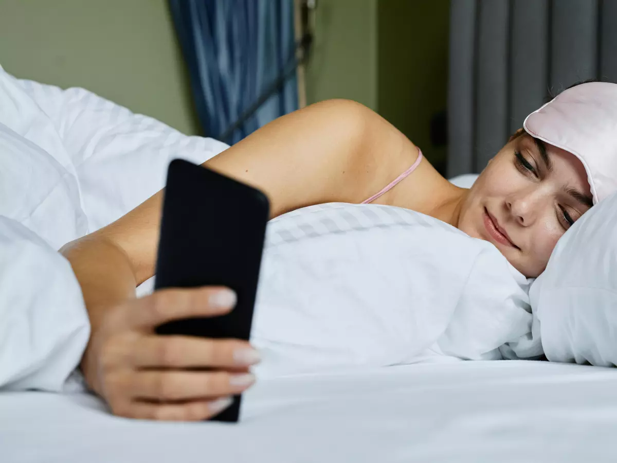 A woman in bed holding a smartphone in her hand, eyes closed.