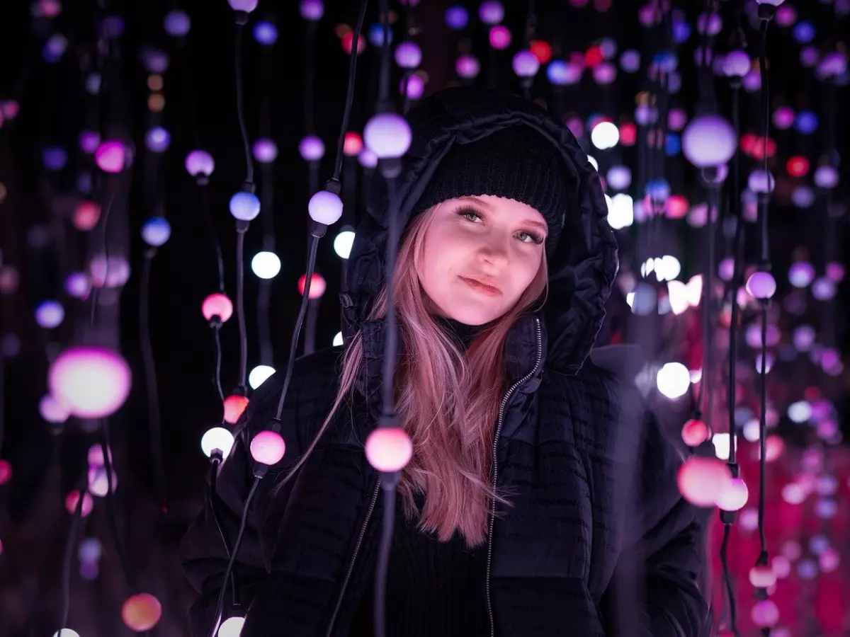 A young woman with long blonde hair stands against a backdrop of colorful lights, seemingly looking at something to the side of the camera.