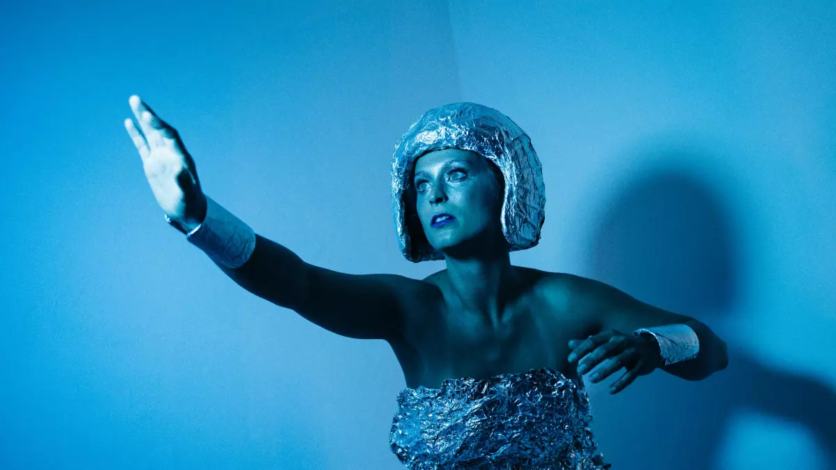 A woman in a silver suit with a helmet stands in a blue room, posing in a robotic-like gesture.