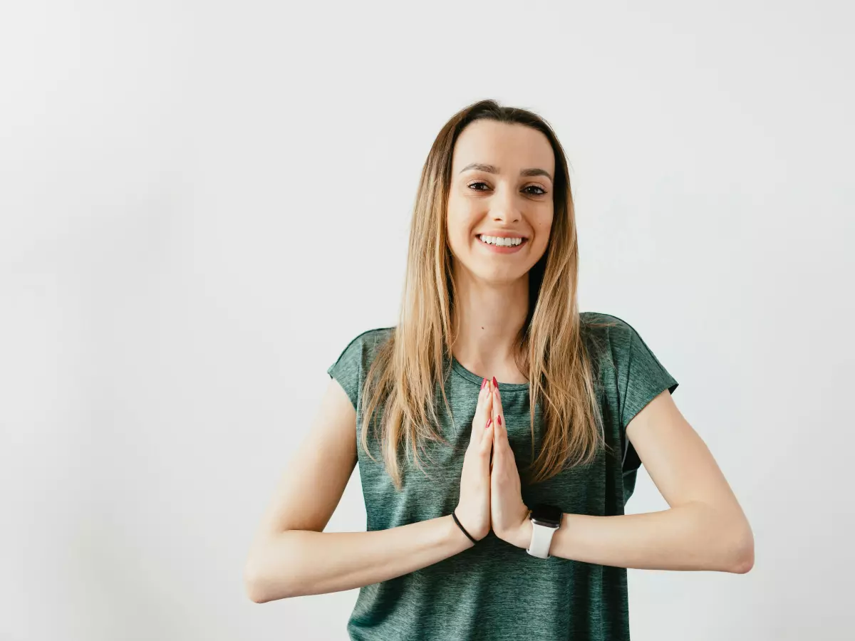 A woman wearing a smartwatch and a green t-shirt, holding her hands together in a prayer pose and smiling at the camera.