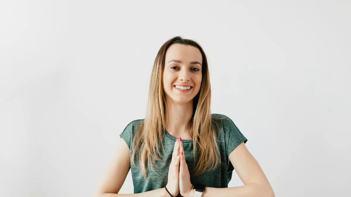 A woman wearing a smartwatch and a green t-shirt, holding her hands together in a prayer pose and smiling at the camera.