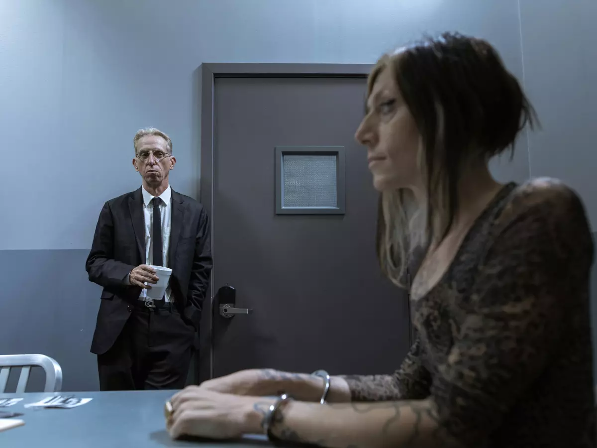 A woman in handcuffs sits at a table, looking worried, while two men in suits stand behind her. The scene takes place in a starkly lit interrogation room.