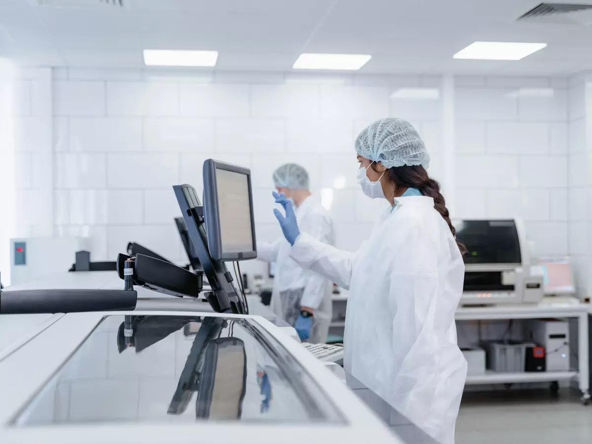 Two people in lab coats are working in a laboratory. One person is looking at a computer screen, while the other is working on a piece of equipment.