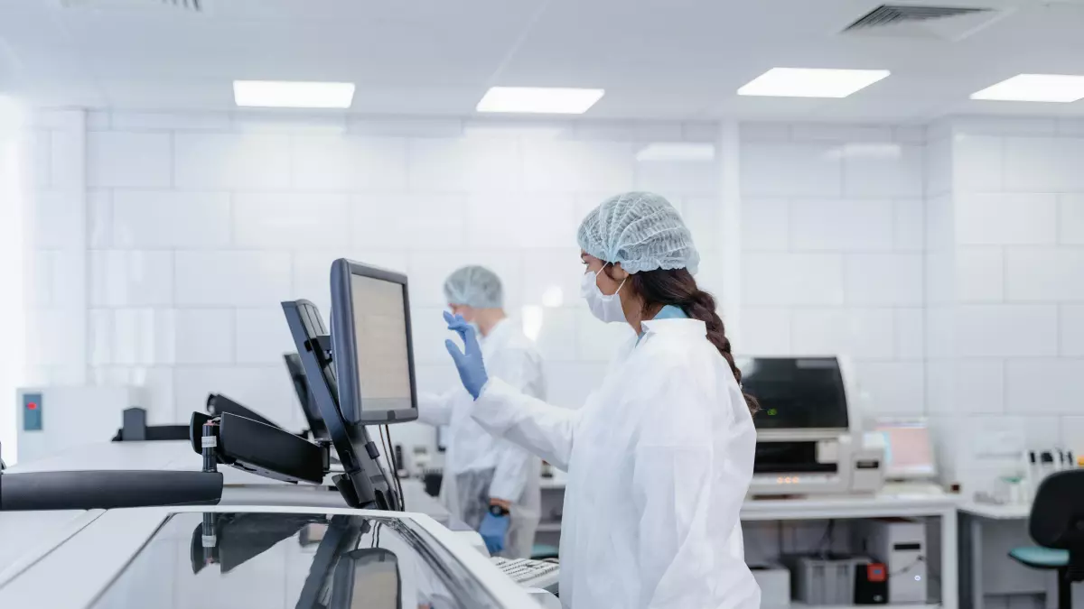 Two people in lab coats are working in a laboratory. One person is looking at a computer screen, while the other is working on a piece of equipment.