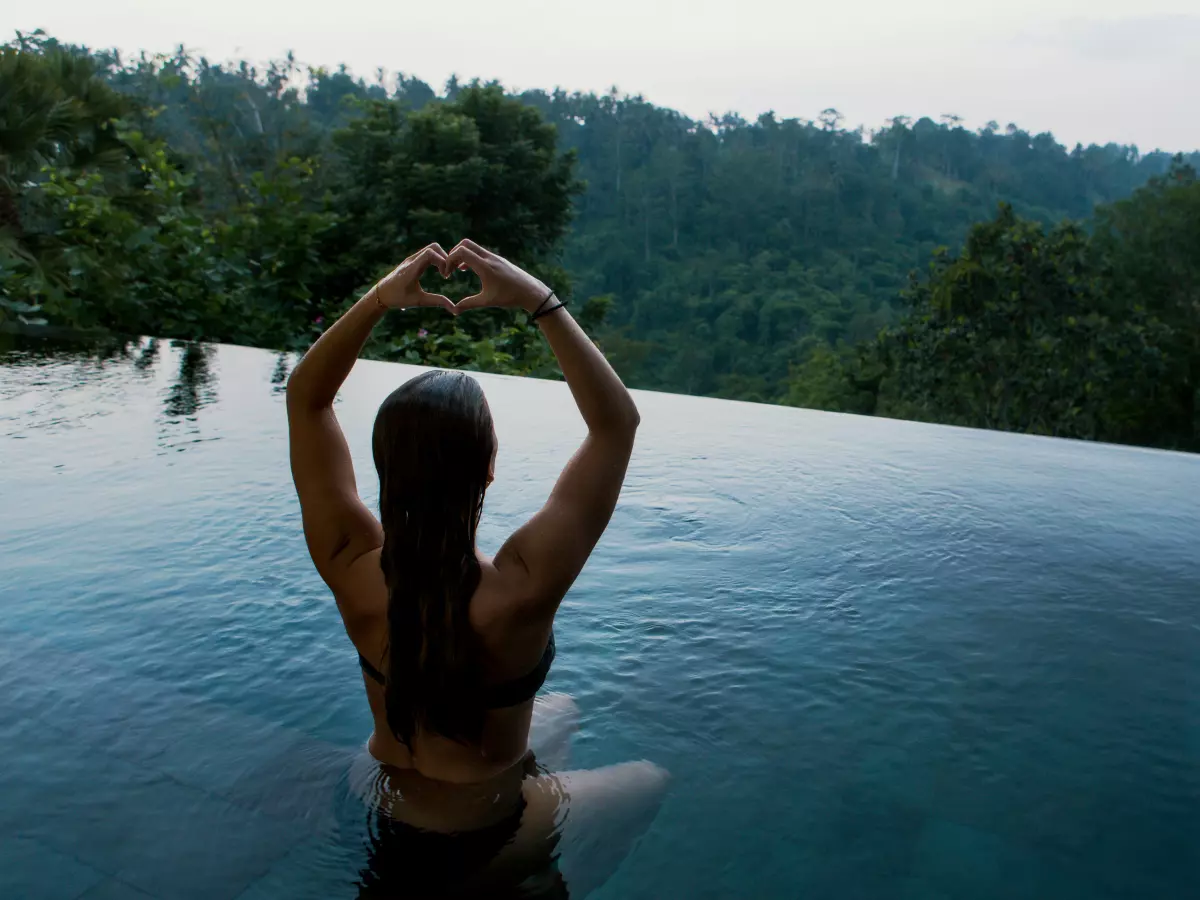 A woman sits on the edge of a pool with her back to the camera, making a heart shape with her hands. The scenery in the background is a green mountain range.