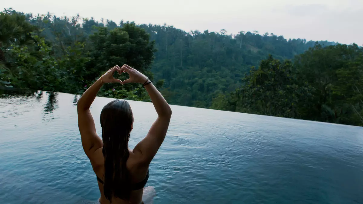 A woman sits on the edge of a pool with her back to the camera, making a heart shape with her hands. The scenery in the background is a green mountain range.