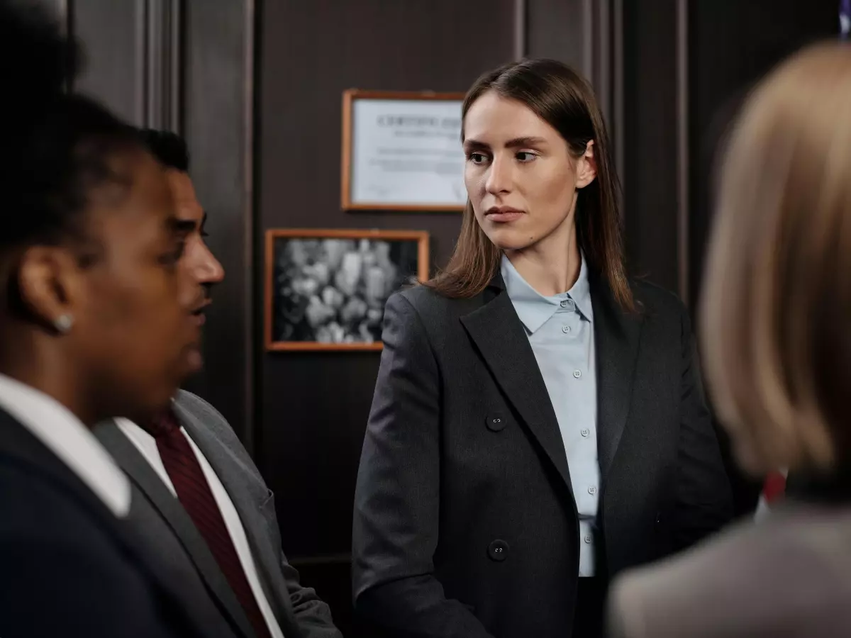 A woman in a black suit stands in a room with two other people. She is looking at the camera and has a serious expression.