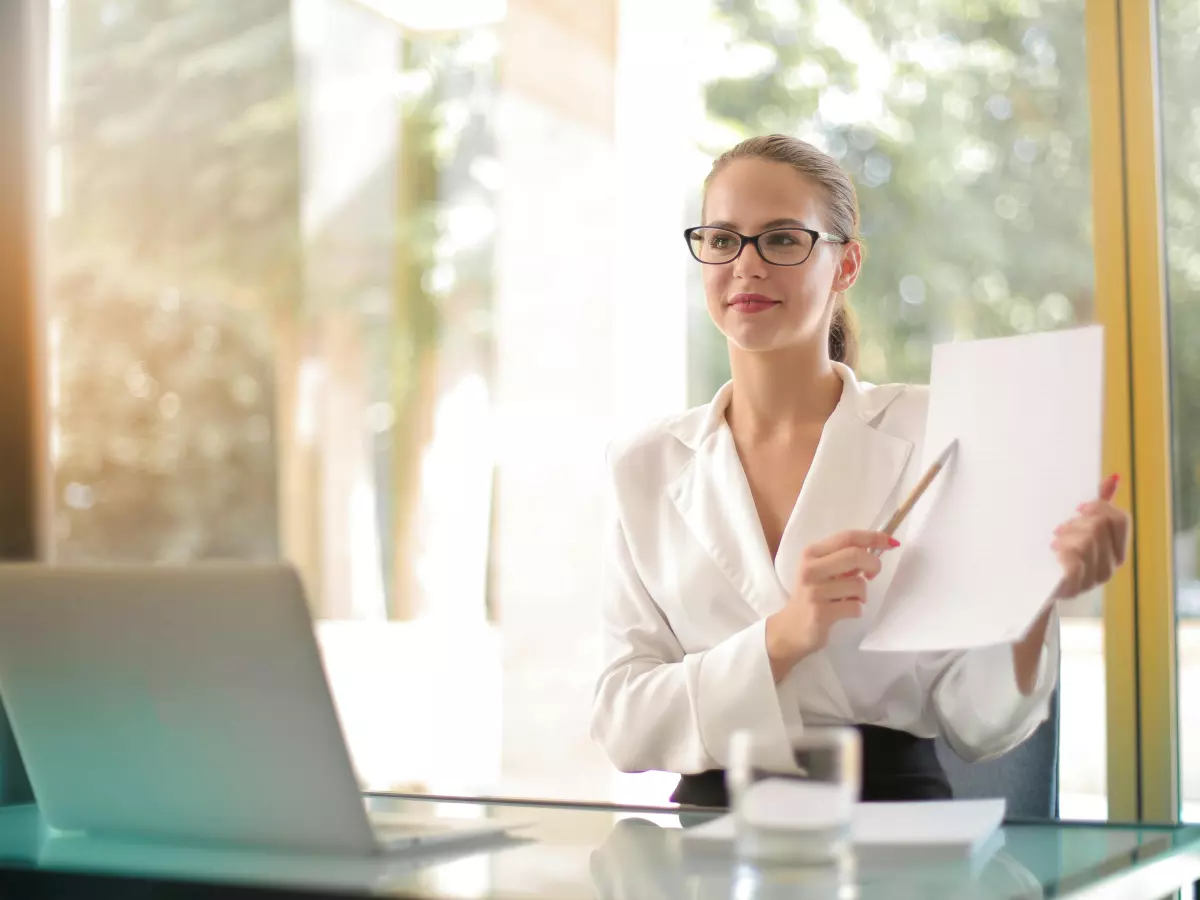 A woman in a white shirt sitting at a desk with a laptop and holding a paper, looking at the camera and talking.