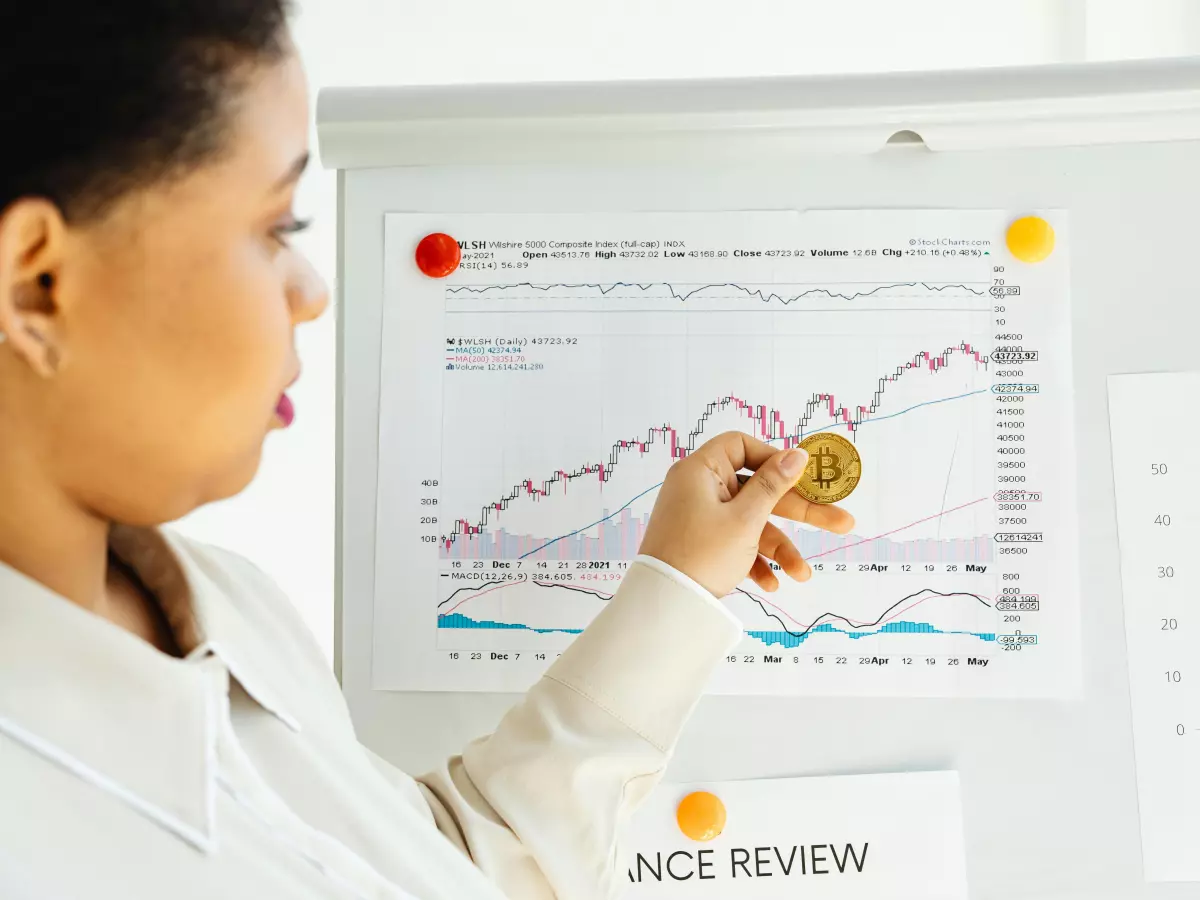 A businesswoman looking at a chart showing an upward trend, holding a Bitcoin coin in her hand. It's a stock image depicting a financial concept.