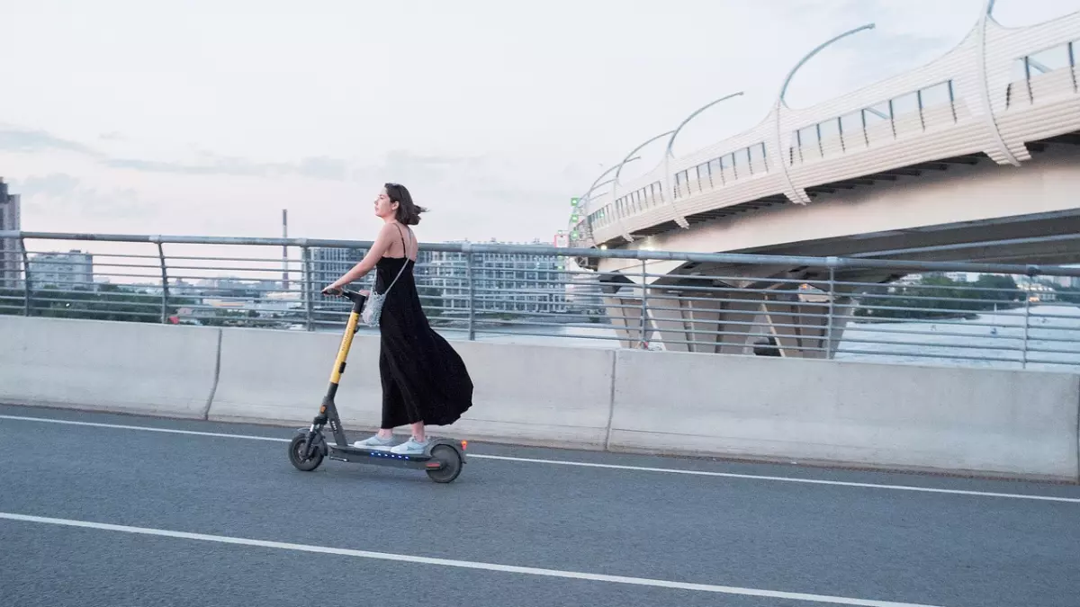 A woman rides an electric scooter on a road with a bridge in the background.