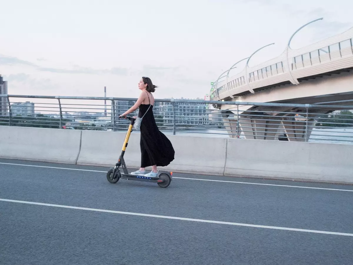 A woman rides an electric scooter on a road with a bridge in the background.