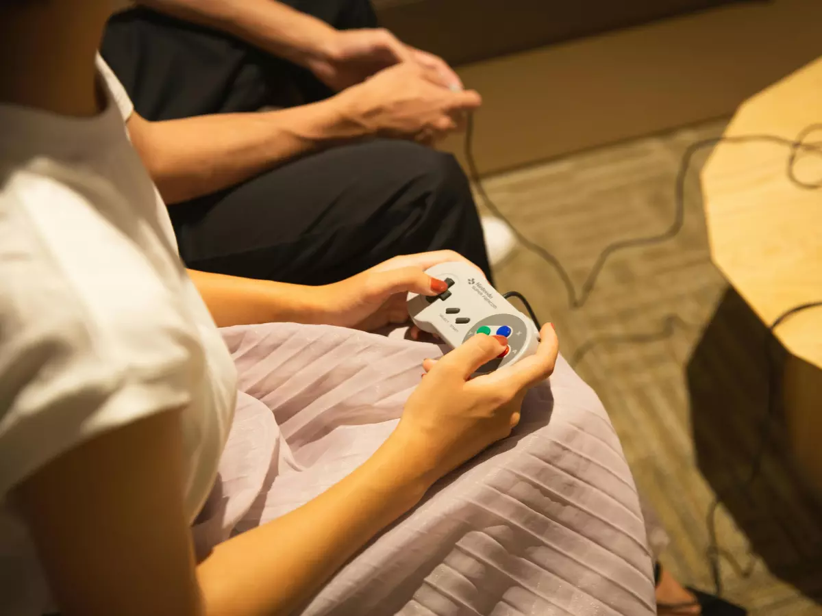 A person playing a retro game with a handheld console, the person is wearing a white shirt and a pink skirt, the background is blurred and the focus is on the person's hands and the console.