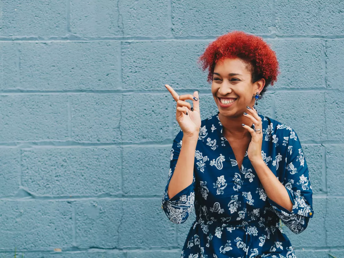 A woman with bright red hair is smiling and pointing to something off-camera.