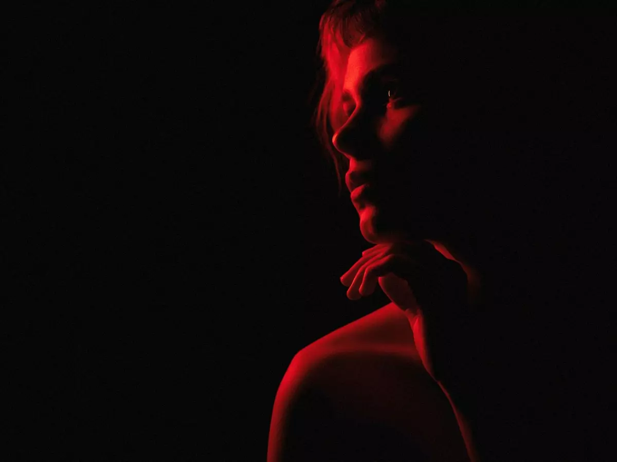 A woman's face is illuminated by a red light, casting shadows on her features. The background is dark. The image focuses on the play of light on her face.