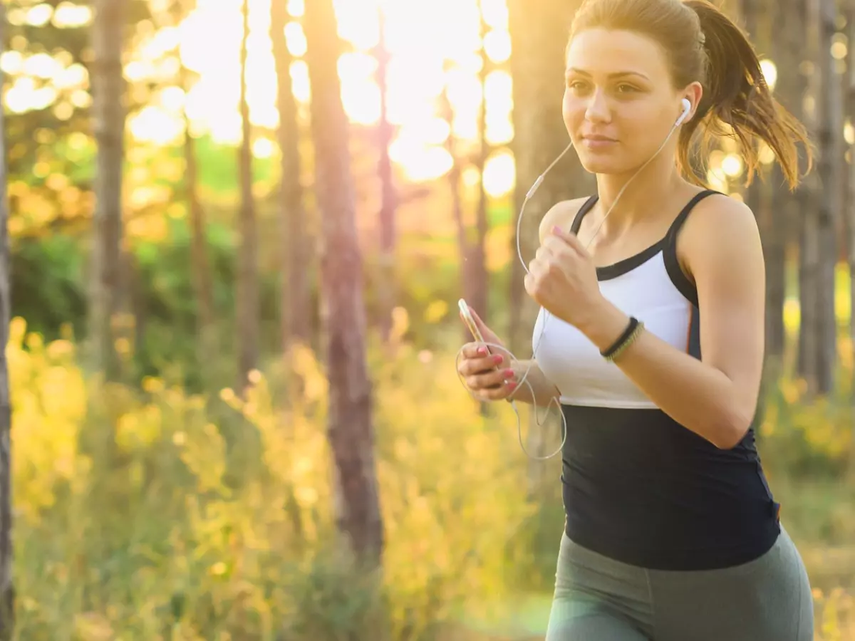 A woman with headphones and a phone in her hand is running through a forest, illuminated by warm sunlight.