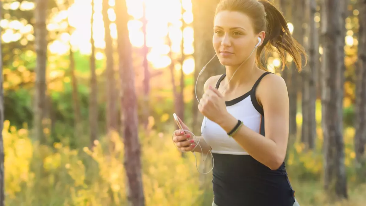 A woman with headphones and a phone in her hand is running through a forest, illuminated by warm sunlight.