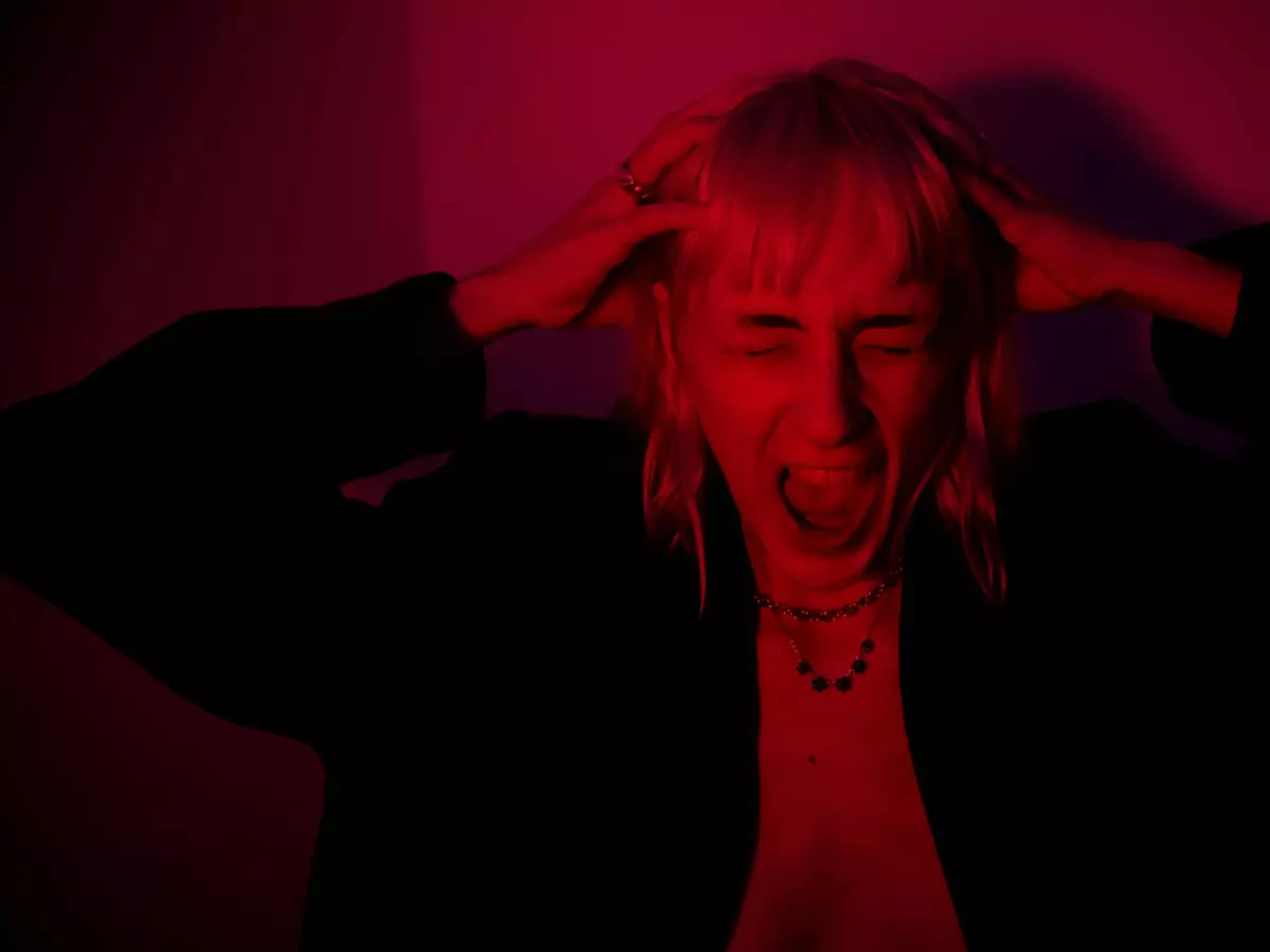 A young woman screams with her hands on her head. The lighting is red and the background is dark.