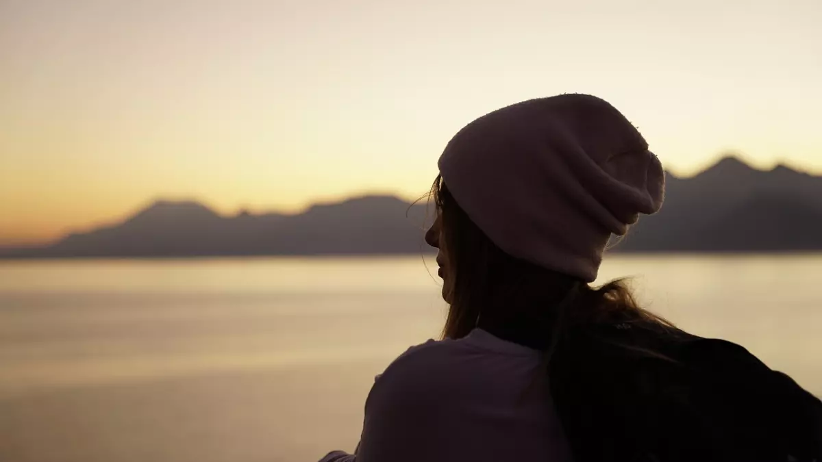 A person with a pink beanie and a backpack stands with their back to the camera, looking out at a sunset over a body of water. Mountains are in the background.