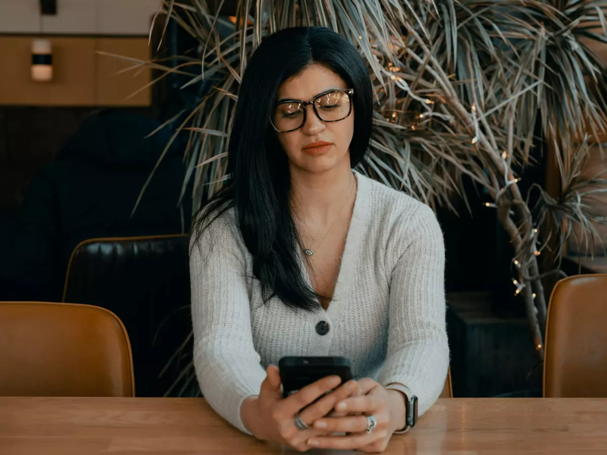 A woman with long dark hair is sitting at a table, looking at her smartphone.