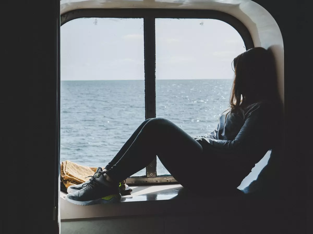 A woman sitting by a window on a cruise ship, looking out at the ocean. The image is taken from inside the ship, with the window frame visible.